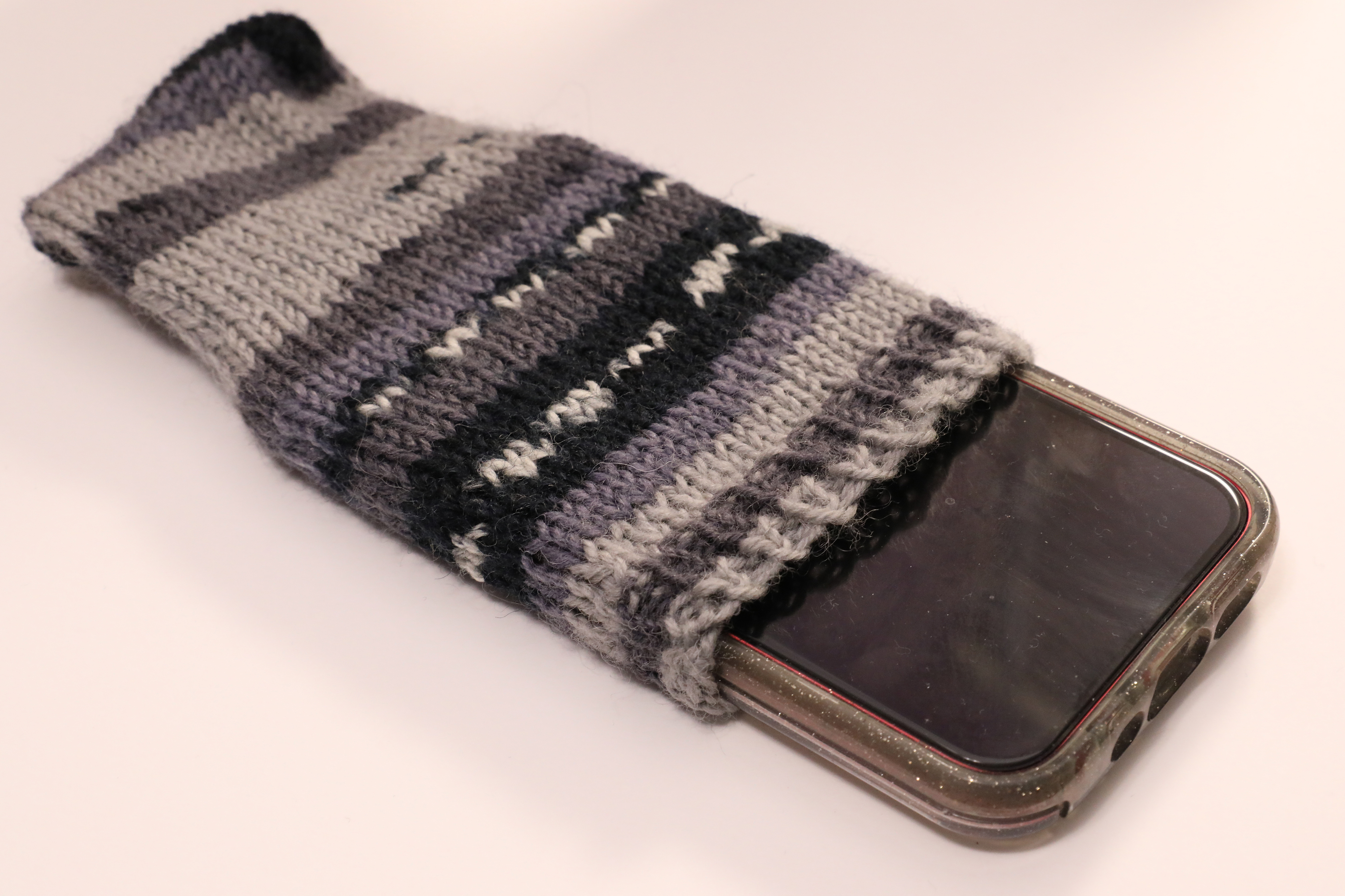 A knit, monochrome (black, white, and shades of grey) cellphone cozy on a white background. A smartphone is inside the cozy, poking out a bit on the open end.